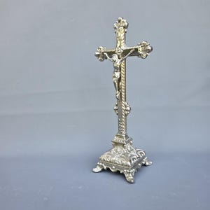 Può includere: Un crocifisso in metallo color argento su una base quadrata decorativa. La croce presenta bordi testurizzati e ornati e una figura centrale di Gesù. La base è dettagliata con intricati motivi a volute e poggia su quattro piccoli piedini.