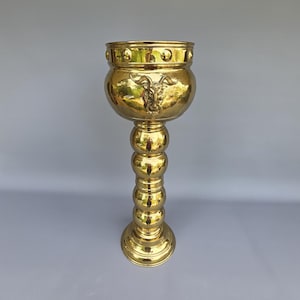 May include: A tall, golden brass planter with a round bowl-shaped top and a decorative goat head emblem. The planter has a series of spherical accents on the stem and a circular base. The planter is suitable for indoor or outdoor use.