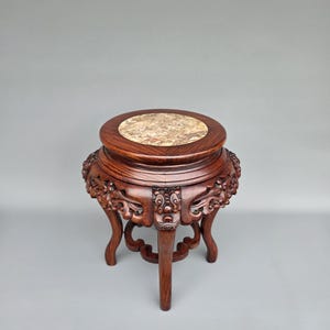 Op de afbeelding: Een kleine, ingewikkeld gesneden houten standaard met een ronde bovenkant en drie poten. De bovenkant heeft een gespikkelde, lichtbruine marmeren inzet. Het donkerbruine hout is gedetailleerd met sierlijke gravures rond de basis en poten.