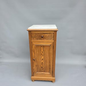 Op de afbeelding: Een kleine houten kast met een marmeren blad. De kast heeft een enkele lade met een decoratief gesneden ontwerp en een deur met een verticaal houtnerfpatroon. De kast is ongeveer 76 cm hoog.