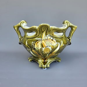 Może przedstawiać: Zielony ceramiczny wazon w stylu Art Nouveau z kwiatowym wzorem. Wazon ma szeroki, otwarty wierzch z uchwytami i podstawą na cokole. Kwiatowy wzór przedstawia żółty i biały irys. Wazon ma około 20 cm szerokości i 15 cm wysokości.