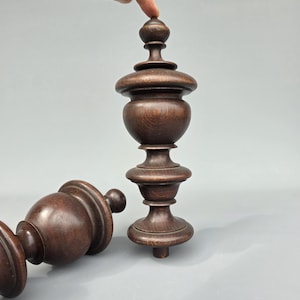May include: Two dark brown wooden decorative finials with a turned design. One finial is upright, and the other is lying on its side. The finials have a bulbous shape with a series of stacked, rounded elements. A finger is touching the top of the upright finial.