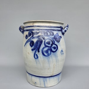 Op de afbeelding: Een witte keramische pot met blauwe bloemmotieven en twee kleine handvatten. De pot heeft een brede opening en een blauwe band rond de basis. Het blauwe bloemmotief staat op de voorkant.