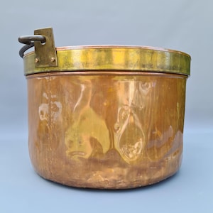 Op de afbeelding: Een koperen pot met een messing rand en handvat. De pot heeft een cilindrische vorm en een aangetaste, verouderde uitstraling. De handgreep is met een messing beugel aan de zijkant bevestigd. De pot is waarschijnlijk een antiek of vintage item.