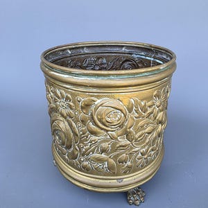 Op de afbeelding: Een vintage messing plantenbak met een bloemenreliëf. De plantenbak heeft een cilindrische vorm met een gedetailleerd patroon van rozen en andere bloemen. Het heeft een verweerde patina en rust op drie kleine, decoratieve voetjes.