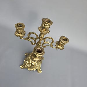 May include: Ornate gold-colored candelabra with four candle holders. The candelabra features a detailed, scrolling design with a decorative base. The candle holders are designed to hold standard-sized candles. The overall design is vintage and elegant.