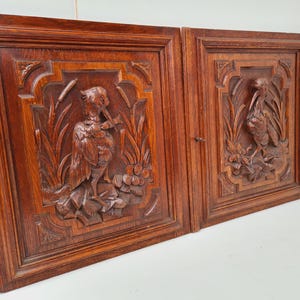 May include: Two antique wooden panels with intricate carvings. Each panel features a detailed relief of a bird amidst foliage. The panels are framed in a rich, dark brown wood, showcasing the craftsmanship of the carvings.