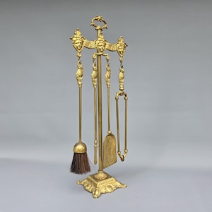 May include: A gold-colored fireplace tool set. The set includes a brush, shovel, poker, and tongs, all suspended from a decorative stand. The stand and tools feature ornate detailing, including sculpted faces and floral motifs. The brush has a dark brown bristle.