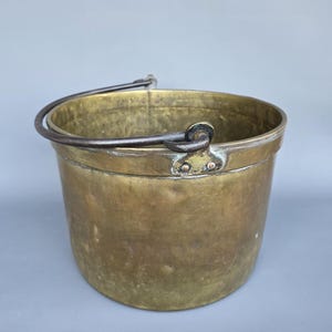 May include: A vintage brass bucket with a dark metal handle. The bucket has a cylindrical shape and a slightly tarnished finish, suggesting age. The handle is attached with rivets. The overall aesthetic is rustic and antique.