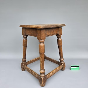 Op de afbeelding: Een kleine, antieke houten kruk met een vierkante bovenkant en vier gedraaide poten. De kruk is gemaakt van lichtbruin hout en heeft een driehoekige basis voor extra stabiliteit. De poten zijn verbonden door een lager frame.