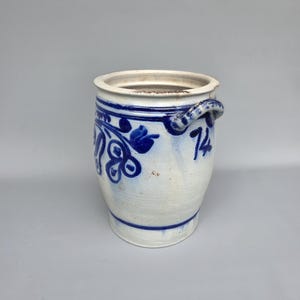 Op de afbeelding: Een witte en blauwe keramische pot met een handvat. De pot heeft blauwe bloemmotieven en het nummer 74. De pot heeft een cilindrische vorm met een licht uitlopende basis. Het handvat is een lus van keramiek.