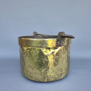 May include: A hammered brass bucket with a dark metal handle. The bucket has a textured surface and a wide, rounded shape. The handle is attached with rivets. The bucket is likely vintage or antique.