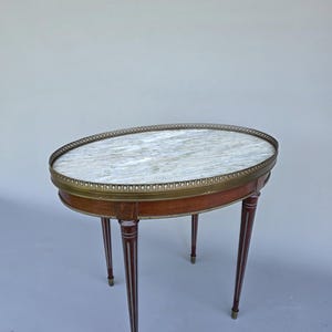 Op de afbeelding: Een ovale bijzettafel met een wit en grijs geaderd marmeren blad, omringd door een decoratieve messing galerijrand. Het houten frame heeft een donker roodbruine afwerking en rust op vier taps toelopende poten met messing voetjes.