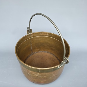 May include: A copper bucket with a rounded body and a metal handle. The bucket has a brass rim and a slightly tarnished appearance. The handle is arched and attached to the sides of the bucket. The bucket's diameter is approximately 25 cm.