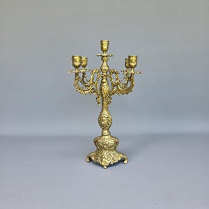 May include: Ornate gold-colored candelabra with five candle holders. The candelabra features a detailed, scrolling design with a decorative base and central column. The candle holders are designed to hold standard-sized candles.