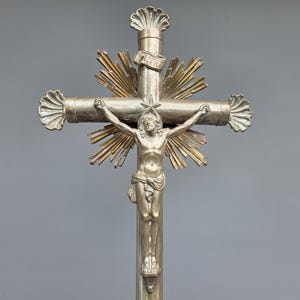 Puede incluir: Crucifijo plateado con una detallada representación de Jesucristo. La cruz presenta detalles ornamentales, incluyendo rayos de sol detrás de la figura y extremos decorativos. Un artículo religioso, adecuado para exhibición. Medidas en centímetros.