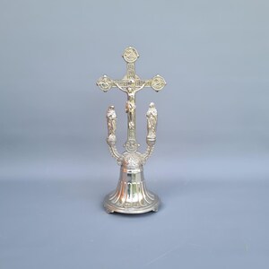 Pode incluir: Um crucifixo de metal prateado sobre uma base alargada e canelada. A cruz apresenta uma figura central de Jesus, ladeada por duas figuras em pé sobre suportes curvos. Quatro medalhões circulares estão posicionados nas extremidades da cruz.