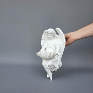 Może przedstawiać: Biała ceramiczna figurka anioła ze szczegółowymi skrzydłami i kwiatową koroną. Anioł trzyma dużą, teksturowaną muszlę. Figurka jest przeznaczona do montażu na ścianie, dodając odrobinę elegancji do każdej przestrzeni.