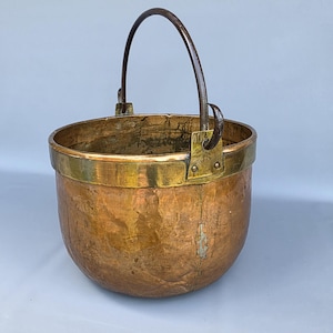 Op de afbeelding: Een koperen en messing emmer met een gebogen metalen handvat. De emmer heeft een afgeronde body met een messing rand en een donker metalen handvat. Het koper heeft een warme, verouderde patina.