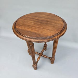 Puede incluir: Una mesa auxiliar redonda de madera con acabado marrón oscuro. El tablero presenta un borde circular con ranuras. La mesa se apoya en cuatro patas estriadas conectadas por un travesaño central en forma de cruz con detalles torneados.