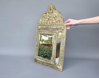 May include: Ornate gold-colored mirror with a rectangular frame and a decorative top. The mirror has intricate carvings and a detailed border. The frame is a warm gold tone, and the mirror reflects a green bush.