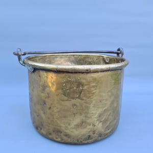 May include: A vintage brass bucket with a rounded body and a metal handle. The bucket has a tarnished, aged appearance, with visible wear and discoloration. The handle is attached to the bucket with metal loops. The bucket is likely used for decorative purposes.