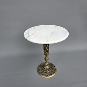 Peut inclure: Petite table d'appoint avec un plateau rond en marbre blanc. La table a un pied et une tige ornés de couleur laiton. La base présente un motif ajouré circulaire. La table mesure environ 61 cm de haut.