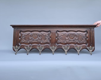 May include: A dark brown wooden wall rack featuring two rectangular panels with intricate floral carvings. The bottom edge has a scalloped design with seven metallic hooks, each adorned with a detailed cherub or angel face motif.