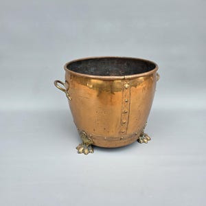 Op de afbeelding: Koperen emmer met messing handgrepen en decoratieve messing klauwpoten. De emmer heeft een geklonken ontwerp en een donker interieur. Het koper heeft een warme, verouderde patina.