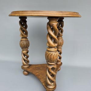 Op de afbeelding: Een kleine, antieke houten tafel met een rond blad en drie ingewikkeld gesneden, spiraalvormig gedraaide poten. De tafel is gemaakt van lichtbruin hout en heeft een klein plankje. De tafel is ongeveer 61 cm hoog.