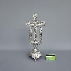 Pode incluir: Um crucifixo prateado com um design detalhado. A cruz apresenta a figura de Jesus e é ladeada por duas estátuas. A base é ornamentada e decorativa, com um padrão floral. O crucifixo está sobre um fundo cinzento.
