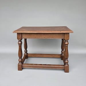 Może przedstawiać: Mały, kwadratowy drewniany stolik w ciepłym brązowym kolorze. Stół ma prostokątny blat wsparty na czterech toczonych nogach i dolnej poprzeczce. Projekt jest klasyczny i solidny, odpowiedni do różnych zastosowań. Wymiary: około 60 cm x 60 cm.