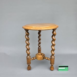 Op de afbeelding: Een ronde houten bijzettafel met een glad bovenblad en drie gedraaide poten. De poten zijn verbonden door een centrale ronde houten voet aan de onderkant. Een standaard creditcard ligt naast de tafel als referentie voor de schaal.