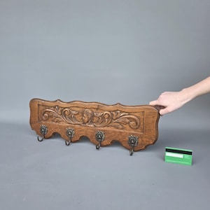 Könnte beinhalten: Ein Vintage-Garderobenständer aus Holz mit kunstvollen Schnitzereien und fünf Metallhaken. Der Ständer hat eine warme braune Farbe und weist ein detailliertes Reliefdesign mit einer zentralen Figur und Scrollmustern auf. Die Haken sind mit Löwenköpfen verziert.
