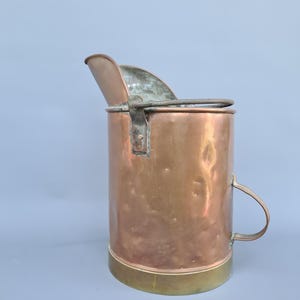 Könnte beinhalten: Ein kupferner Kohleeimer mit Messingfuß und Griff. Der Eimer hat eine gebogene Oberseite und einen Klappdeckel. Das Kupfer hat einen warmen, rötlich-braunen Farbton, und der Messingfuß fügt einen kontrastierenden Goldton hinzu.