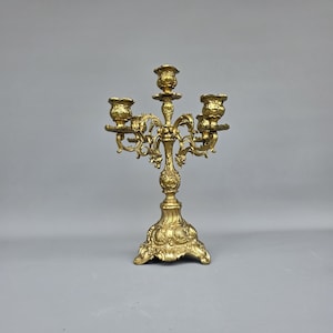 May include: A gold-colored candelabra with five candle holders. The candelabra has an ornate design with floral and scrollwork details. The base is wide and decorative, and the overall style is vintage.