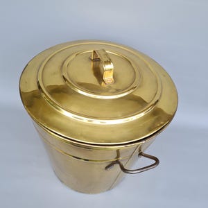 Könnte beinhalten: Ein Vintage-Messing-Eimer mit Deckel und Griff. Der Eimer hat eine zylindrische Form mit einem abgerundeten Deckel und einem kleinen Griff oben. Ein gebogener Griff ist an der Seite des Eimers befestigt.