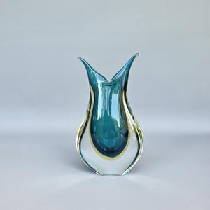 Könnte beinhalten: Eine klare Glasvase mit tealfarbenem Innenraum und einem gelb-goldenen Band. Die Vase hat eine tulpenförmige Form mit einem ausgestellten Oberteil. Das Glas ist glatt und reflektierend, mit glänzendem Finish. Die Vase steht vor einem hellgrauen Hintergrund.