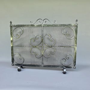 May include: A rectangular metal fireplace screen featuring a fine mesh center panel framed by a dark metallic border. The design includes decorative scrollwork accents in each corner and a central symmetrical pattern, supported by two small feet.