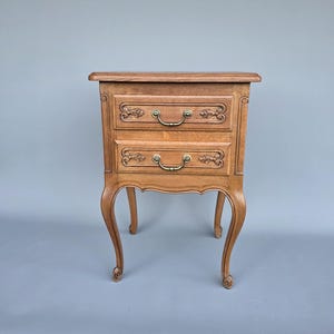 May include: A light brown, two-drawer nightstand with ornate carvings and curved legs. The drawers have brass-colored handles. The nightstand is made of wood and has a classic, vintage design.