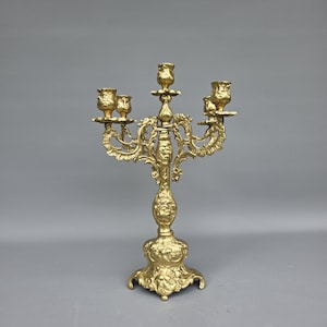 May include: A gold-colored metal candelabra featuring five candle holders arranged on ornate, scrolling arms. The central stem and base are decorated with intricate relief carvings, including facial motifs and floral patterns throughout the structure.
