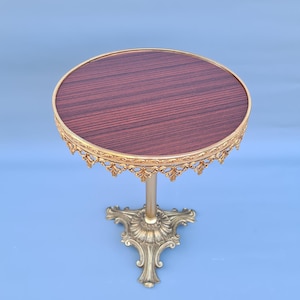 Può includere: Un tavolino laterale piccolo e ornato con un piano rotondo con un motivo a venature di legno scuro. Il tavolo ha un bordo decorativo color oro e una base a piedistallo abbinata. Il tavolo è alto circa 61 cm.