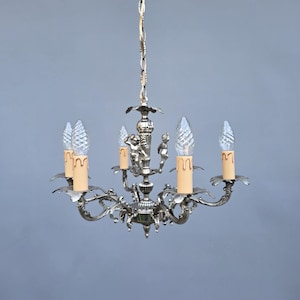 May include: Silver chandelier with five candle-style lights. The fixture has ornate, curved arms and decorative elements, including small figures. The bulbs are clear and shaped like flames. The chandelier hangs from a chain.