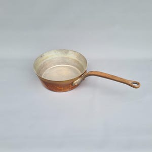 Peut inclure: Une casserole en cuivre avec un long manche en bois. L'intérieur de la casserole est de couleur argentée et l'extérieur présente une patine brun-rougeâtre. La poignée est fixée avec un petit rivet métallique. La casserole est conçue pour la cuisine.