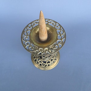 Op de afbeelding: Een vintage messing wierookhouder met een opengewerkt ontwerp. De houder heeft een centrale beker om wierook in te bewaren en een houten kegel. Het messing heeft een verouderde patina en het ontwerp is sierlijk.