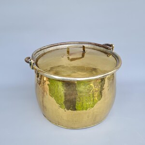 Puede incluir: Una brillante olla de cocina antigua de latón con tapa. La olla tiene forma redondeada con un asa a cada lado y una tapa con una pequeña asa. El latón tiene un color dorado cálido, que refleja la luz.