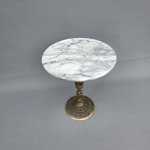 May include: A round side table featuring a white marble top with grey veining. The table is supported by a single, ornate, metallic bronze-toned pedestal base with intricate openwork detailing and a circular, decorative footed base.