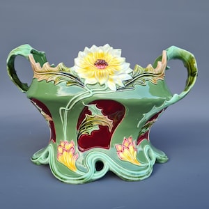 May include: A ceramic jardiniere featuring a sage green base with deep red recessed panels. The vessel is adorned with raised, sculpted yellow and white flower blossoms, green leaves, and two curved handles on the sides with a scalloped base.