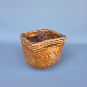 以下が含まれることがあります： 温かみのある土色をした、風化した正方形の陶器鉢。 鉢はわずかにテクスチャ加工された表面と、湾曲した本体の周りに水平な溝があります。 内部には使用感が見られます。 鉢はくすんだ青色の背景に置かれています。