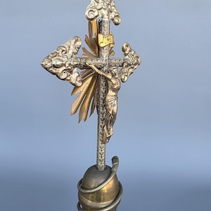 May include: A detailed, antique crucifix with a gold-toned Christ figure. The cross is silver with gold accents, including a sunburst design behind the figure. The base is a brass-colored sphere with a coiled design.